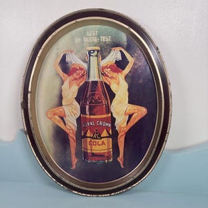 May include: An oval metal serving tray featuring a vintage advertisement for Royal Crown Cola. The center displays a brown glass bottle with the text RC, Royal Crown, and Cola. Two figures in white dresses flank the bottle, with the text Best By Taste-Test printed above. The tray has a dark brown rim.