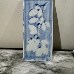 May include: Rectangular ceramic dish with a blue and white floral design. The dish features a blue background with vertical brushstrokes and white leaf and stem details. The edges are slightly raised.