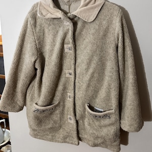 May include: A beige, button-down jacket with a corduroy collar and two front pockets. The jacket has three-quarter length sleeves and is made of a soft, textured material. The pockets have a decorative embroidered design.