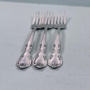 May include: A set of three silver-toned metal forks featuring ornate, embossed scrollwork detailing along the handles. Each fork has four tines and a polished finish, with the decorative pattern extending from the base to the neck of the handle.