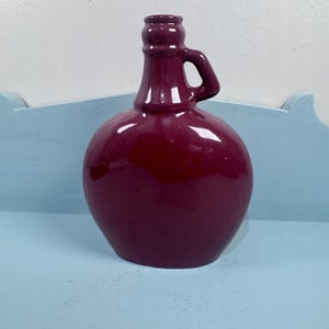 May include: A deep magenta ceramic jug with a rounded, flattened body and a narrow neck. The vessel features a single small handle attached to the side of the neck, with a smooth, glossy finish reflecting light across its curved surface.