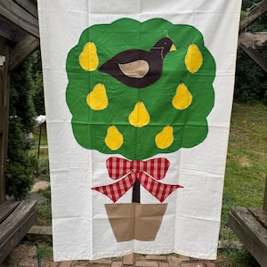 May include: A white fabric banner featuring a green tree with yellow pears and a brown bird. A red and white plaid bow adorns the tree trunk, which is planted in a brown pot. The banner is hanging outdoors.
