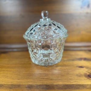 May include: A clear, vintage glass candy dish with a lid. The dish has a textured, diamond-like pattern on the sides and lid. The lid is topped with a small, rounded handle. The dish sits on a wooden surface.