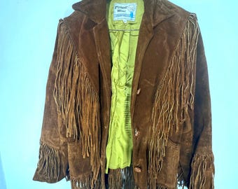 Puede incluir: Chaqueta de ante marrón con largos flecos en las mangas, el pecho y la espalda. La chaqueta tiene cuello y botones en la parte delantera. El forro interior es de color verde brillante. La etiqueta dice "Pioneer Wear".