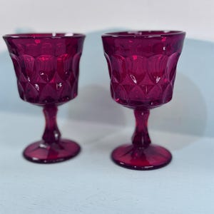 May include: Two deep magenta colored glass goblets featuring a textured diamond pattern on the bowl, a short stem, and a circular base. The translucent glassware has a faceted surface design that catches the light, creating a uniform appearance.