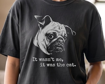 May include: A charcoal grey short-sleeve t-shirt featuring a white graphic illustration of a pug's face. Below the dog, the text reads, "It wasn't me, it was the cat." The shirt has a crew neckline and a relaxed, loose-fitting silhouette.