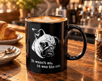 May include: A black ceramic mug featuring a white line-art illustration of a pug's face and the text "It wasn't me, it was the cat." The mug is filled with coffee topped with latte art and steam rising from the surface.