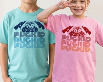 May include: A light blue t-shirt and a pink t-shirt, each featuring a graphic of a pug face above the text PUGKID repeated three times in a stacked, linear font. The blue shirt has dark blue text, while the pink shirt has red and orange text.