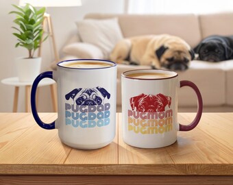 May include: Two white ceramic mugs with coloured handles and rims. The left mug has a blue handle and features a pug illustration with the text PUG DAD in blue. The right mug has a maroon handle and features a pug illustration with the text PUG MOM in red.
