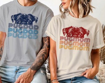 May include: A light grey t-shirt featuring a dark blue pug illustration and the text PUGDAD in blue retro-style lines, alongside a cream t-shirt with a dark red pug illustration and the text PUGMOM in red, orange, and yellow retro-style lines.