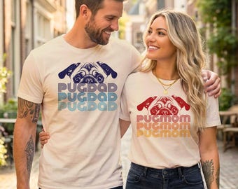 May include: Two cream-coloured short-sleeve t-shirts. One shirt features a blue pug illustration with the text PUGDAD in a retro font. The other shirt features a red pug illustration with the text PUGMOM in a retro font. Both shirts have a crew neckline.