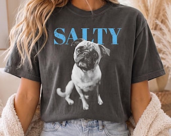 May include: A charcoal grey short-sleeve t-shirt featuring a black and white photographic print of a pug dog sitting down. Above the dog, the word SALTY is printed in a distressed light blue font. The shirt has a crew neckline and a relaxed fit.