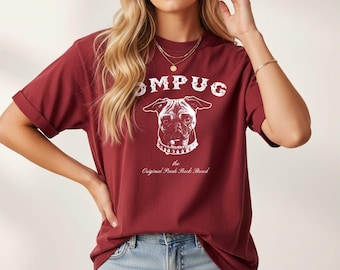 May include: A maroon short-sleeve t-shirt featuring a white graphic of a pug wearing a spiked collar. The text reads "POMPUG" above the dog and "the Original Punk Rock Breed" below the dog. The shirt has a crew neckline and rolled sleeves.