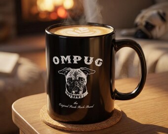 May include: A black ceramic mug filled with steaming coffee featuring a white graphic of a pug wearing a spiked collar. The text OMPUG and the phrase the Original Punk Rock Breed are printed in white on the front of the mug, which sits on a cork coaster.