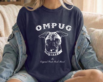 May include: A navy blue t-shirt featuring a white graphic of a pug wearing a spiked collar. The text OMPUG is printed above the dog, and the text the Original Punk Rock Breed is printed below the dog in a white, distressed, vintage-style font.
