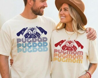 May include: Two off-white t-shirts featuring pug dog illustrations. The left shirt displays a dark blue pug face with the text PUGDAD in stacked blue and light blue lettering. The right shirt shows a red pug face with PUGMOM in red and orange.