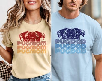 May include: A cream-coloured t-shirt featuring a red pug graphic and the text PUGMOM in a retro font, alongside a light blue t-shirt featuring a dark blue pug graphic and the text PUGDAD in a retro font, both with short sleeves and crew necklines.