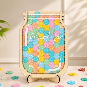 May include: A light-colored wooden jar-shaped reward container featuring the text "Reward Jar" in white script. The interior is filled with numerous small, colorful circular tokens, each embossed with a smiling face, in shades of blue, pink, yellow, orange, and green.