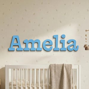 May include: Blue wooden letters spelling "Amelia" are mounted on a wall above a white crib. The letters are in a bold, sans-serif font. The wall has a light beige floral pattern. A beige blanket is draped over the crib.