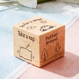 May include: A wooden cube with engraved illustrations and text. The cube features drawings of a car, sun, clouds, a pillow, a tree, and a person walking. Text includes "Go for a drive", "Take a nap", and "Putter Around".