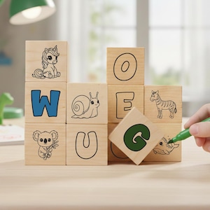 May include: Wooden alphabet blocks with animal illustrations and letters. A green pen is held near a block with the letter "G". Other blocks show a unicorn, snail, zebra, koala, and the letters W, O, E, and U. A green desk lamp is in the background.