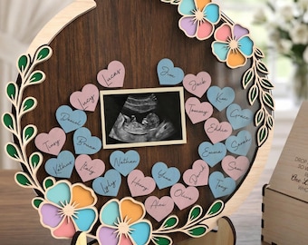 May include: A wooden baby shower guest book with a dark brown circular frame, floral accents, and heart-shaped name tags. The frame holds a sonogram photo and a box with the text "Welcome, please sign a heart".
