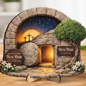 Può includere: Una scena pasquale decorativa in legno raffigurante il sepolcro vuoto con un arco in pietra, una grande pietra e un piccolo albero. La scena comprende un cielo notturno con una luna piena e delle croci. Il testo sulla scena recita "He is Risen".