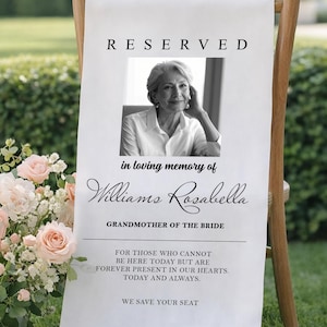 May include: A white fabric sign with the word "RESERVED" at the top. Below is a black and white photo of a woman, with text that reads "in loving memory of Williams Rosabella, Grandmother of the Bride." The sign is on a wooden chair.