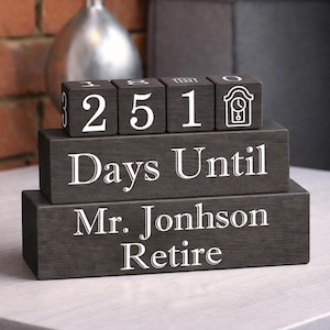 May include: A dark grey wooden countdown block set. The top blocks show the numbers 2, 5, 1, and a clock icon. The middle block reads "Days Until". The bottom block says "Mr. Jonhson Retire".