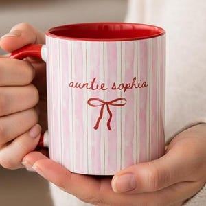 K&ouml;nnte beinhalten: Eine Keramik-Tasse mit rotem Innenraum und Henkel, mit einem rosa-wei&szlig; gestreiften Muster. Die Tasse hat den Text "auntie sophia" und ein rotes Schleifendesign. Die Tasse wird von einer Person gehalten.