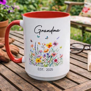 May include: A white ceramic mug with a red interior and handle. The mug features the word "Grandma" in script, along with a floral design and butterflies. The text "EST. 2025" is also present. The mug is on a wooden table.