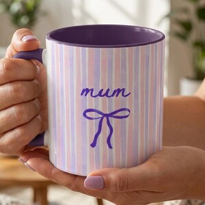 May include: A white ceramic mug with a purple interior and handle. The exterior features vertical stripes in pink, white, and lavender. The word "mum" is written in cursive, with a purple bow below.