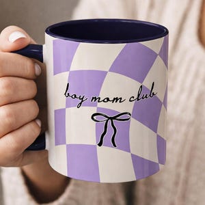Op de afbeelding: Een witte keramische mok met een paars en wit geblokt patroon. De mok heeft een donkerblauwe binnenkant en een handvat. De woorden "boy mom club" staan in zwart script op de mok, met een zwarte strik eronder.