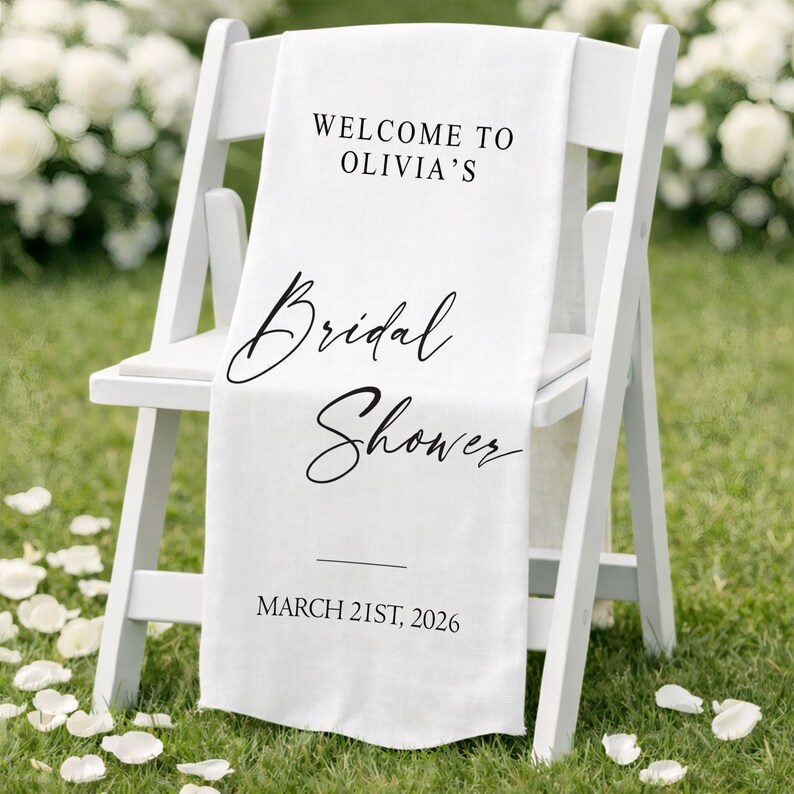 May include: A white chair with a white fabric draped over it. The fabric has the text "WELCOME TO OLIVIA'S Bridal Shower MARCH 21ST, 2026" in black script. The chair is on a grassy surface with white flower petals.
