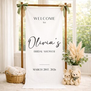 May include: A white fabric banner with the text "WELCOME to Olivia's BRIDAL SHOWER MARCH 21ST, 2026" is displayed on a wooden frame. Decorative elements include a basket of rolled towels, a vase of pampas grass, and two stuffed animals.