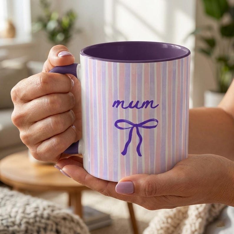 May include: A white ceramic mug with a purple interior and handle. The exterior features vertical stripes in pink, white, and lavender. The word "mum" is written in cursive, with a purple bow below.