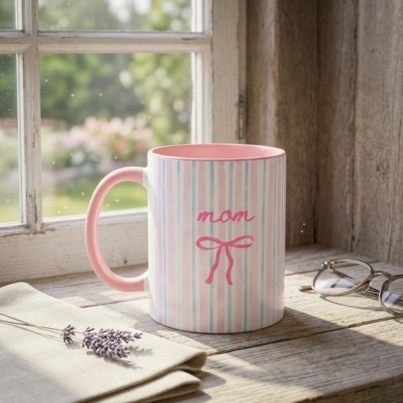 May include: A ceramic mug with a pink handle and rim, featuring vertical stripes in pink, white, and light blue. The word "mom" is written in pink script, with a pink bow below. The mug sits on a wooden surface.