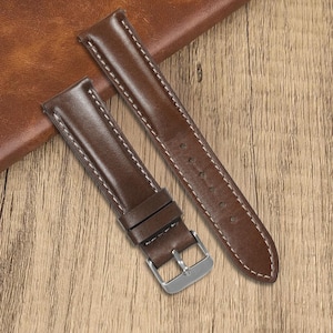 Peut inclure: Deux bracelets de montre en cuir marron avec des coutures blanches et une boucle argentée. Les bracelets sont présentés sur une surface en bois, mettant en valeur la texture et le design des accessoires de montre.