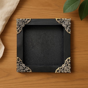 May include: A square, black shadow box with ornate, gold-colored corner accents. The interior is black, and the box is set on a wooden surface. The decorative corners add a vintage or antique aesthetic.