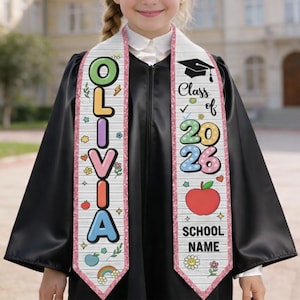 May include: A graduation sash featuring the name "OLIVIA" in colourful letters on one side and "Class of 2026" on the other. The sash has a white background with pink trim and decorative elements including flowers and a rainbow.