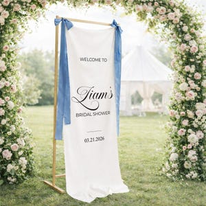 May include: A white fabric banner with the text "WELCOME TO Liam's BRIDAL SHOWER 03.21.2026" in black script. The banner is suspended from a wooden frame and adorned with blue ribbons. The background features a floral arch and a white tent.
