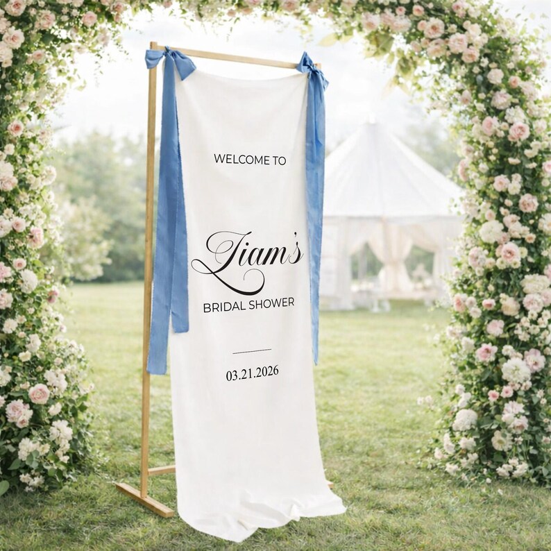 May include: A white fabric banner with the text "WELCOME TO Liam's BRIDAL SHOWER 03.21.2026" in black script. The banner is suspended from a wooden frame and adorned with blue ribbons. The background features a floral arch and a white tent.