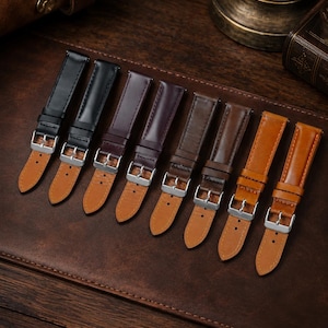 May include: A collection of eight leather watch straps arranged in a row, featuring a gradient of colors from black to dark brown, medium brown, and tan. Each strap includes a silver-toned metal buckle and a light tan leather underside lining.