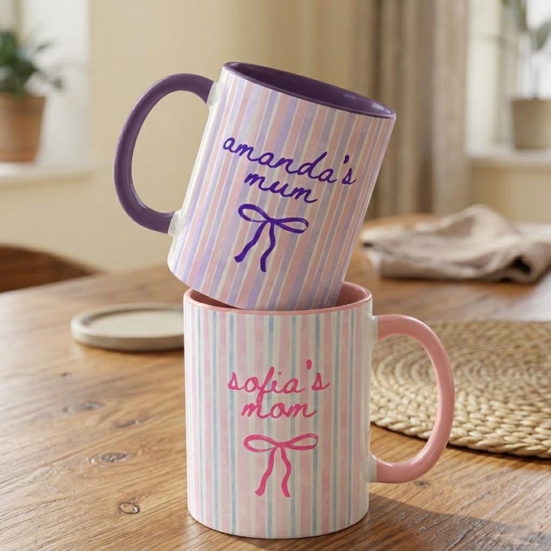 May include: Two ceramic mugs with vertical striped patterns in pink, blue, and white. One mug has a purple handle and the text "amanda's mum" with a bow. The other mug has a pink handle and the text "sofia's mom" with a bow.