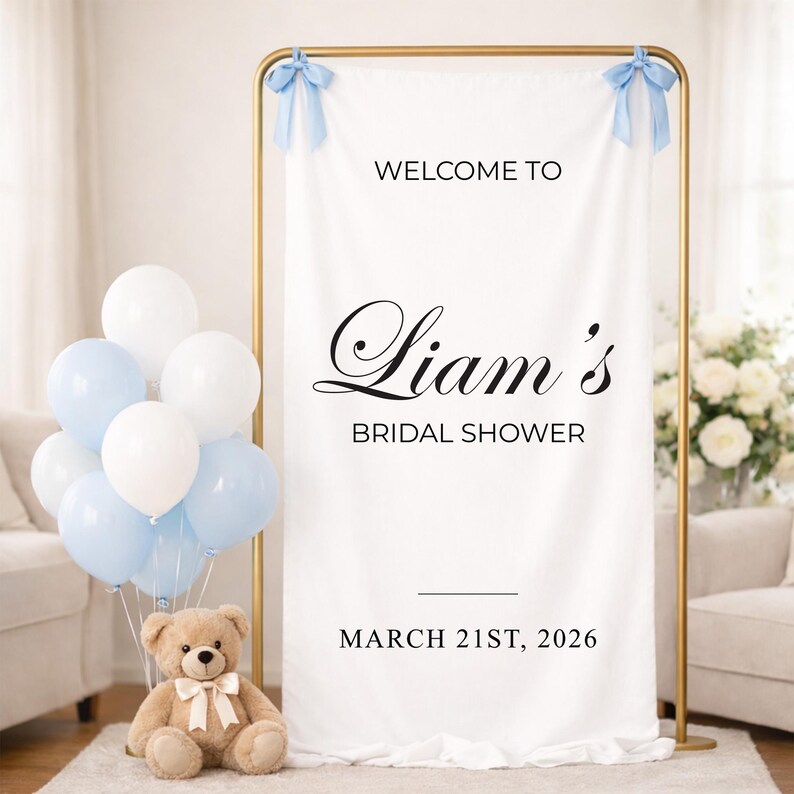 May include: A white fabric banner with the text "Welcome to Liam's Bridal Shower" and the date March 21st, 2026. The banner is held by a gold frame and decorated with blue bows. A cluster of white and light blue balloons and a teddy bear are in front.