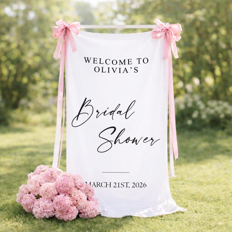 May include: A white fabric sign with the text "WELCOME TO OLIVIA'S Bridal Shower" in black script. The sign is adorned with pink bows and ribbons. A bouquet of pink flowers sits at the base of the sign, with the date "MARCH 21ST, 2026" at the bottom.