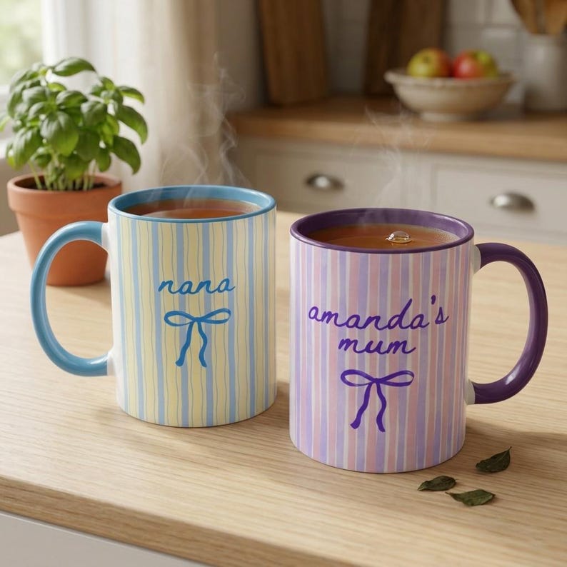 May include: Two ceramic mugs with tea, one with blue and yellow stripes and the word "nana", the other with purple and pink stripes and the words "amanda's mum". Both mugs have a bow design.