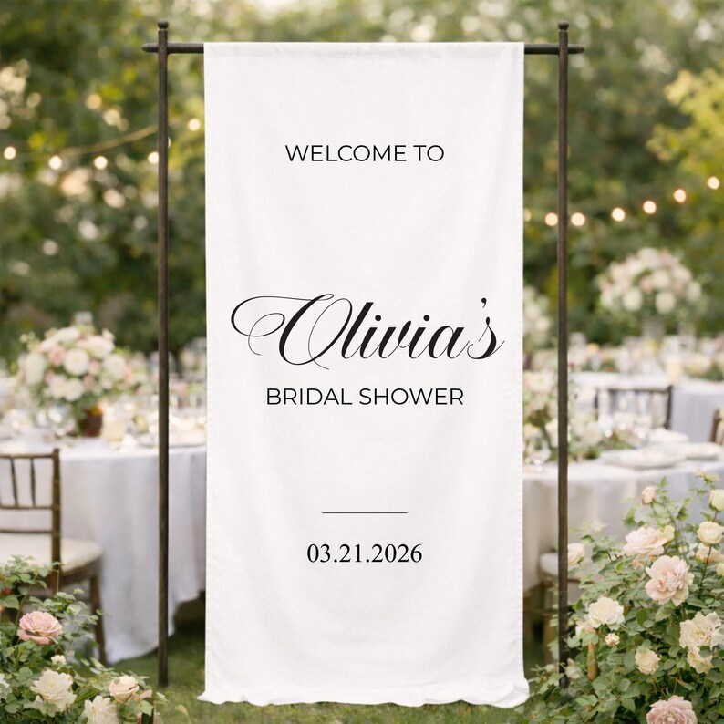 May include: A white banner with black text that reads "WELCOME TO Olivia's BRIDAL SHOWER" and the date 03.21.2026. The banner is hanging on a dark metal stand, with a blurred outdoor background.