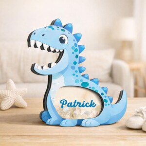 May include: A light blue dinosaur-shaped money box with the name "Patrick" in script. The money box has a large opening for coins and is decorated with darker blue spots and spikes. It is on a wooden surface.