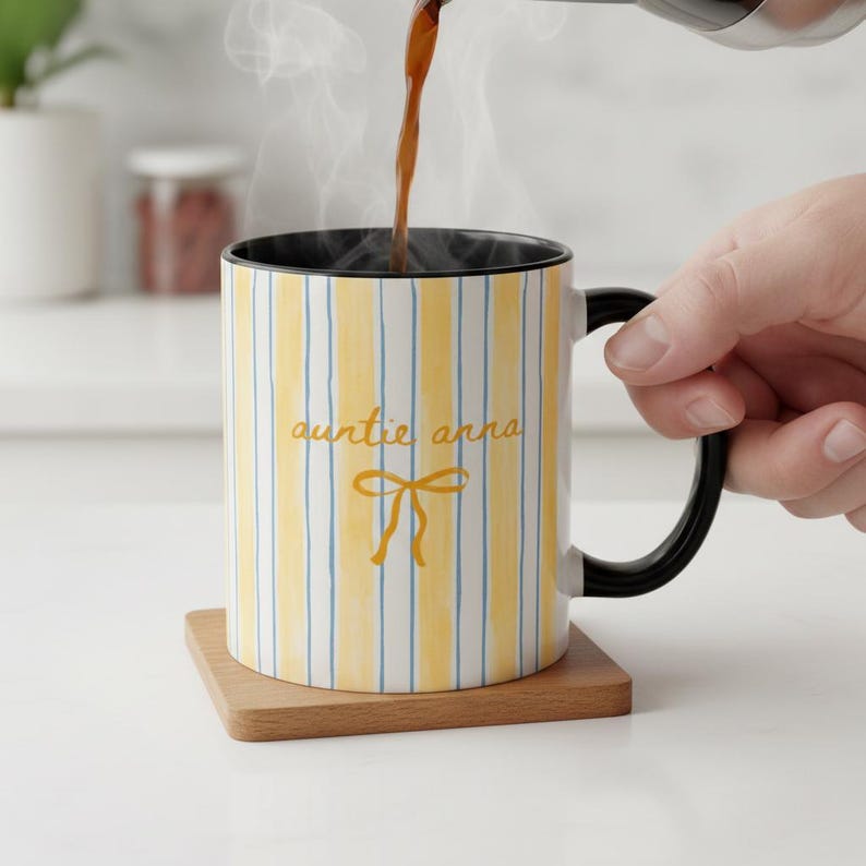 K&ouml;nnte beinhalten: Eine wei&szlig;e Keramiktasse mit gelben vertikalen Streifen und blauen Akzenten. Die Tasse hat einen schwarzen Innenraum und Griff. Auf der Vorderseite stehen die Worte "auntie anna" zusammen mit einer gelben Schleife. Die Tasse steht auf einem Holzuntersetzer.